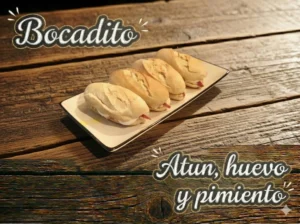 Tres bocadillos en un plato, con atún, huevo y pimiento, sobre una mesa de madera.