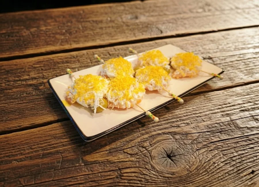 Bandeja con cinco brochetas de aperitivos cubiertos de queso rallado sobre una mesa de madera.